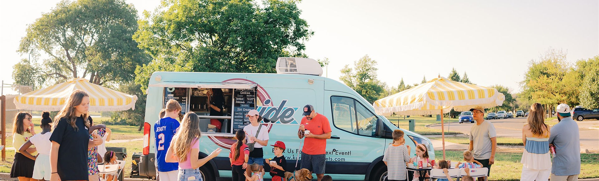 Families lining up at the mint-green Kurbside Sweets truck under yellow striped umbrellas at a Lubbock park